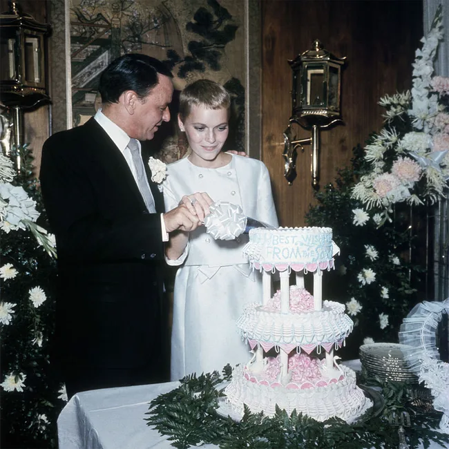 A couple in formal attire cutting a multi-tiered wedding cake surrounded by flowers.