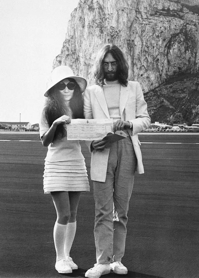 A couple in white attire holding a document, standing on tarmac with a rocky cliff in the background.