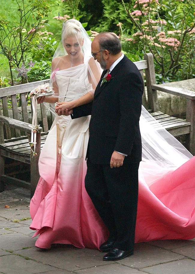 A bride in a white and pink gown walks with an older man in a black suit, surrounded by greenery.