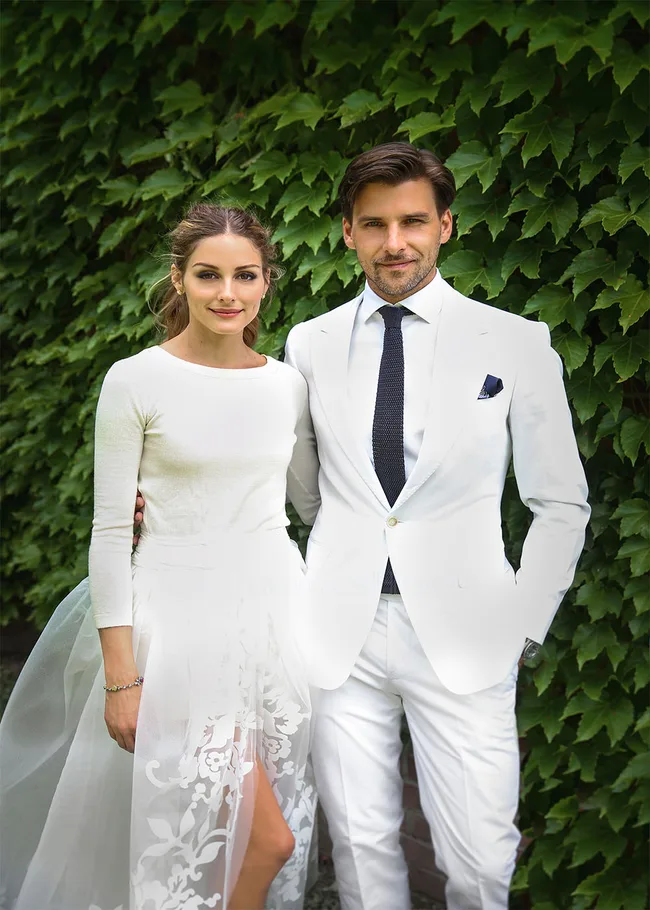 A couple in white formal attire stands smiling against a backdrop of green ivy leaves.