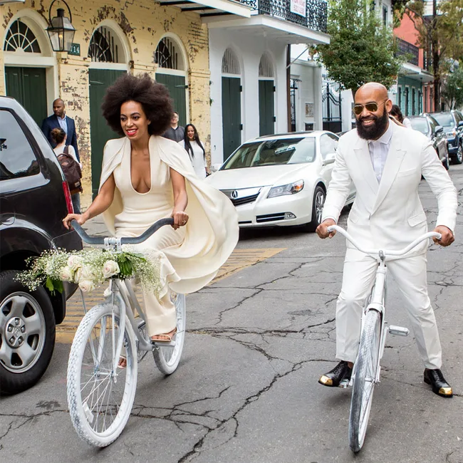 A couple in white outfits cycling together on a street; the woman&rsquo;s bike is decorated with white flowers.