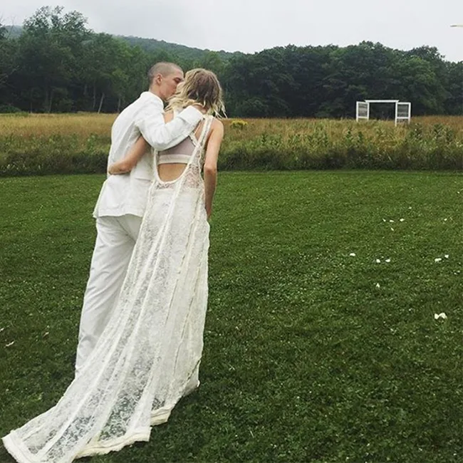 Couple in white, with the woman in a lace dress, standing in a green field with trees and a white structure in the background.