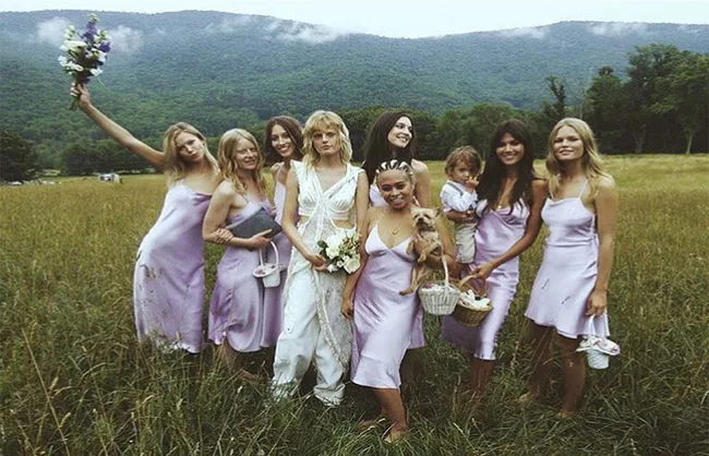 Bridal party in lavender dresses poses in a grassy field with mountains in the background, one holding a bouquet.