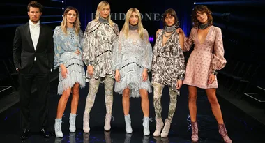 Models on a runway wearing various fashion outfits, including dresses and suits, with a dark background and lights.