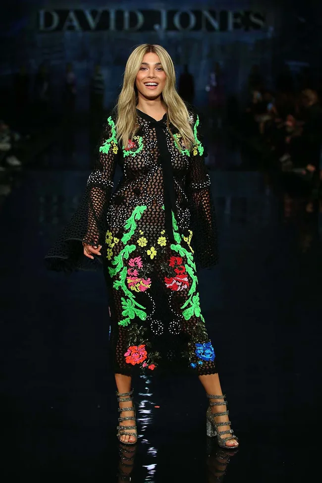 Model on runway in a black floral dress with colorful embroidery, smiling. David Jones sign in the background.