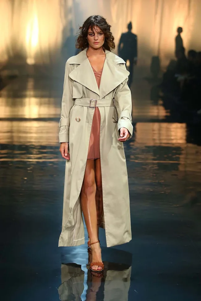 Model on runway in beige trench coat and dress, with background silhouettes on a reflective surface.
