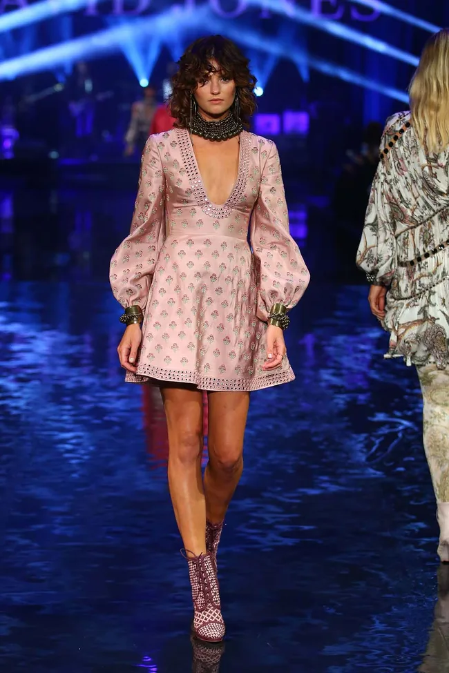 Model in floral pink dress with puff sleeves on runway, blue lighting; fashion show atmosphere.