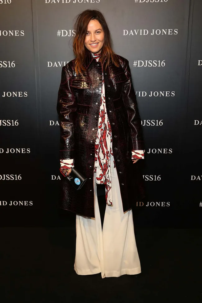 A person in a dark textured coat and white pants at a David Jones event backdrop with "#DJSS16".