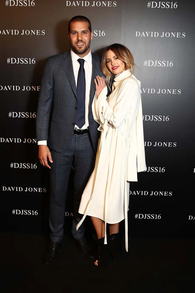 A man in a suit and a woman in a white coat pose together at a David Jones event backdrop.