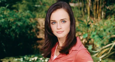 A woman with brown hair in a red top stands outdoors with blurred greenery in the background.