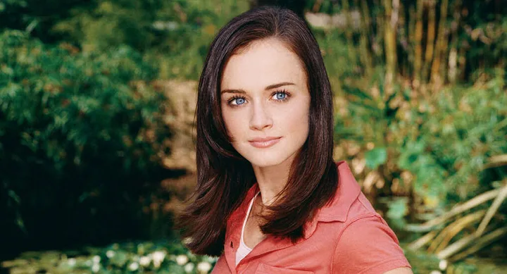 A woman with brown hair in a red top stands outdoors with blurred greenery in the background.