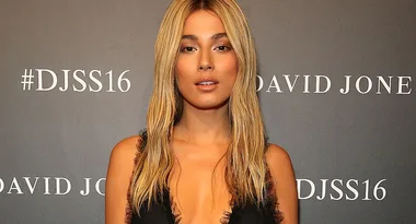 A woman with long blonde hair in a black dress poses in front of a wall with "#DJSS16" and "David Jones" text.