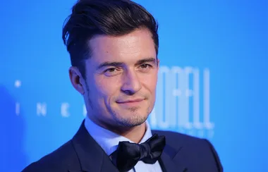 A person in a suit and bow tie smiles against a blue background at an event.