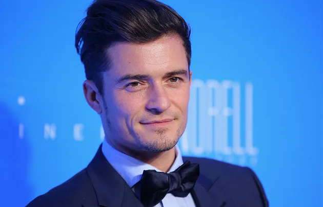 A person in a suit and bow tie smiles against a blue background at an event.