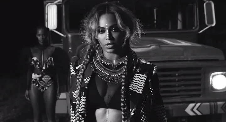 A woman with braids and patterned attire stands in front of a truck at night, with another person in the background.