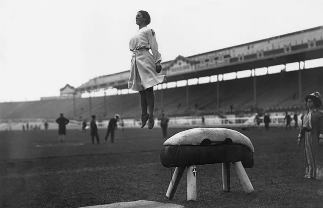 A gymnast in mid-air above a vaulting horse at an early 1900s outdoor competition, with spectators in the background.
