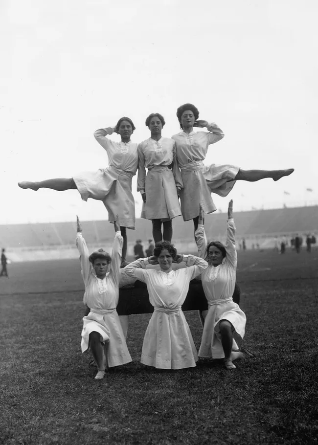 Six women form a human pyramid, performing gymnastics on a field in early 1900s attire.