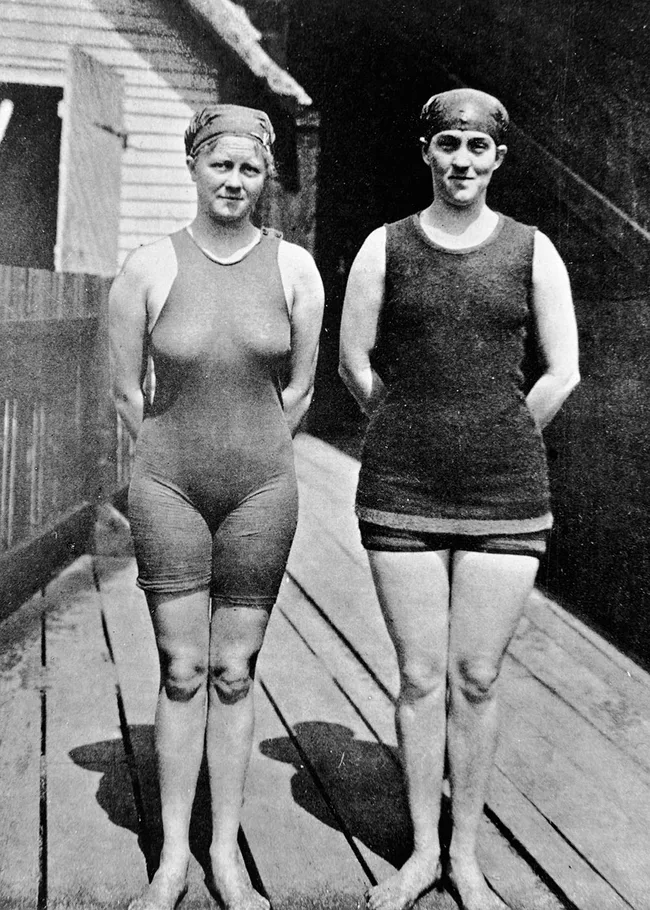 Two women in early 1900s swimsuits and swim caps stand on a wooden deck by a building.