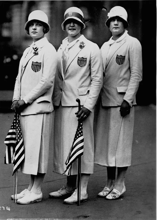 Three women in 1920s athletic outfits with USA emblems, holding American flags, wearing cloche hats.