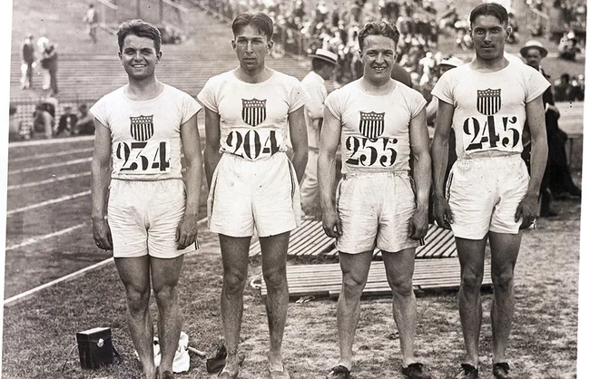 Four athletes wearing USA uniforms, numbered 254, 204, 255, and 245, stand on a track field with spectators behind.