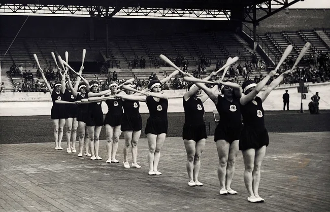 Group of women exercising with clubs in sync on a wooden platform inside a stadium.