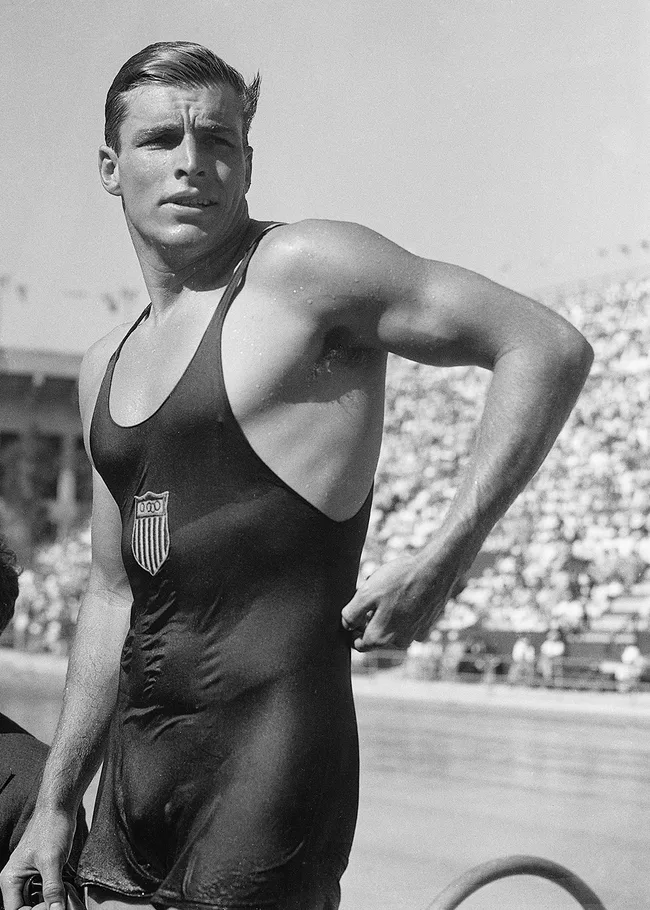 A male athlete in a USA swimsuit stands poolside, looking away, at a sporting event.