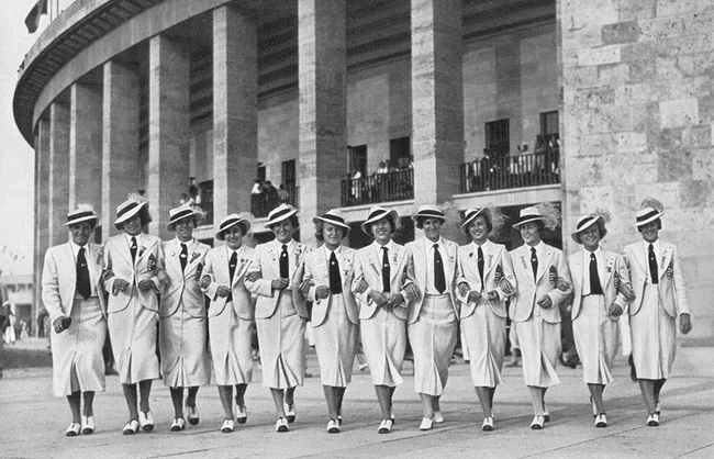 A group of women in matching white suits and hats walk arm-in-arm outside a large stone building.