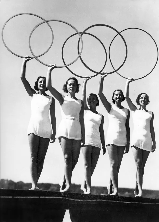 Five women in swimsuits holding large hoops, standing in a line against a clear sky, evoking the Olympic rings.