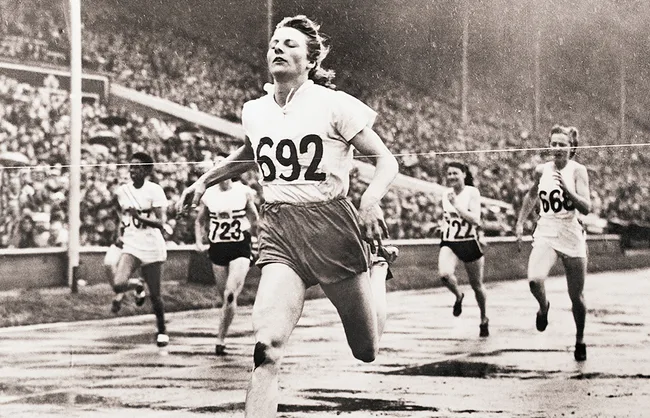 Black and white photo of a female athlete crossing the finish line in a race, with others behind her in a stadium.