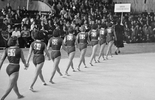 Gymnasts in numbered leotards walk in line at a USSR event, with a sign holder in front and crowd in background.
