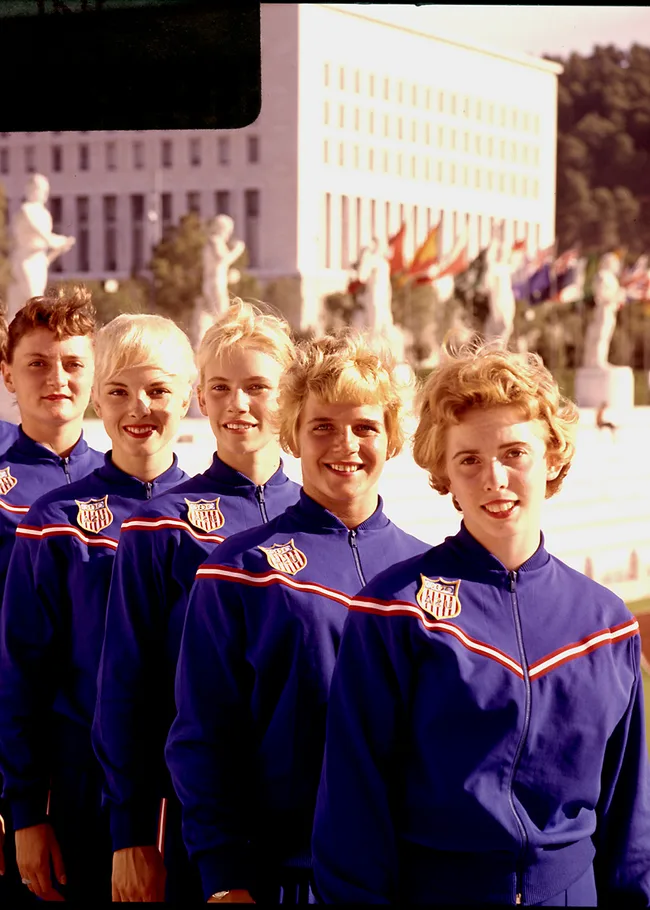 A line of five women in blue USA tracksuits posing in front of stadium statues, likely at an Olympic event.