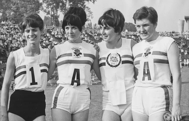 Four female athletes in 1967 England sports uniforms, smiling on a track field.