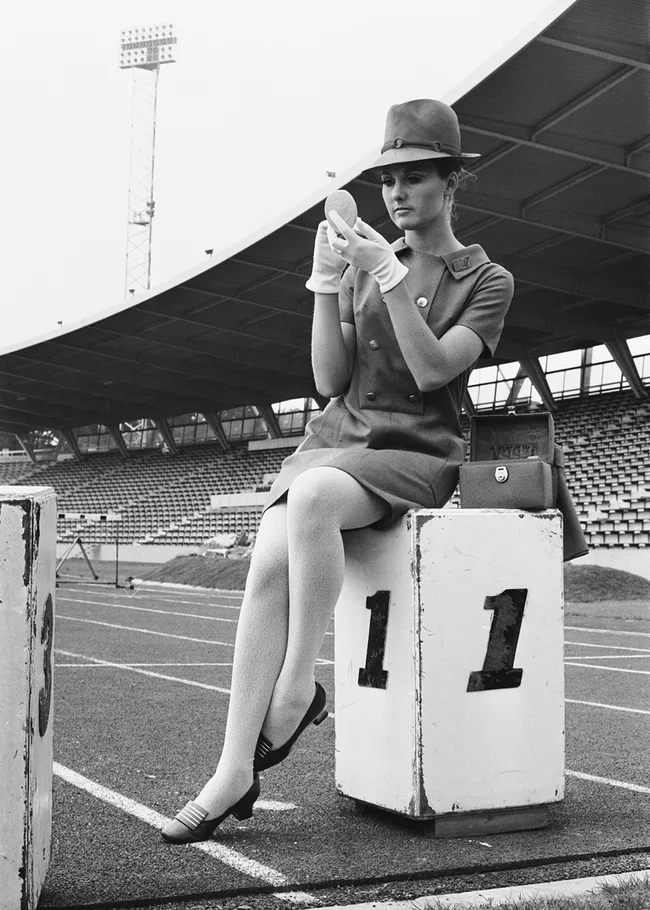 Woman in a vintage outfit with gloves and hat, sitting on a podium at an empty stadium, holding a compact mirror.