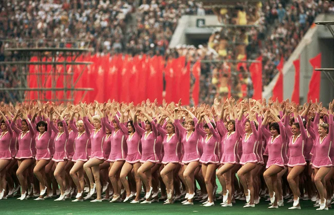 A large group of women in pink outfits perform a synchronized dance at a stadium event.