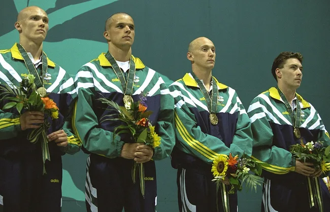Australian men's swimming team on podium with medals and flowers in green and yellow jackets at an event.
