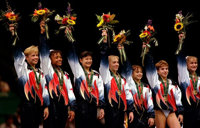 The Magnificent Seven gymnasts in team jackets raising flowers, each wearing a gold medal.