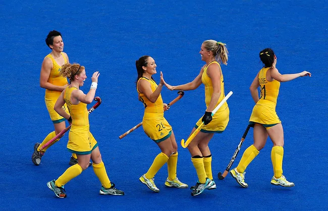 Australian women's field hockey team celebrating on a blue field, wearing yellow uniforms, holding sticks.