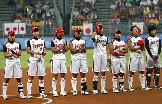 Seven softball players from Japan's team stand in a line on the field, wearing uniforms and caps, facing forward.