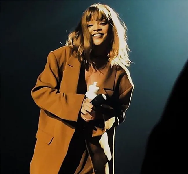 Singer on stage smiling, wearing an oversized brown coat, illuminated by spotlight.