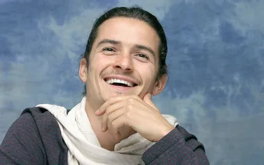 A person smiling with long hair, wearing a scarf and a striped shirt, against a cloudy blue background.
