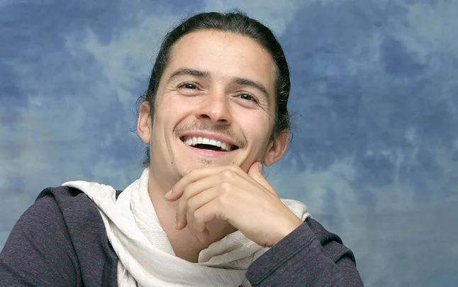 A person smiling with long hair, wearing a scarf and a striped shirt, against a cloudy blue background.