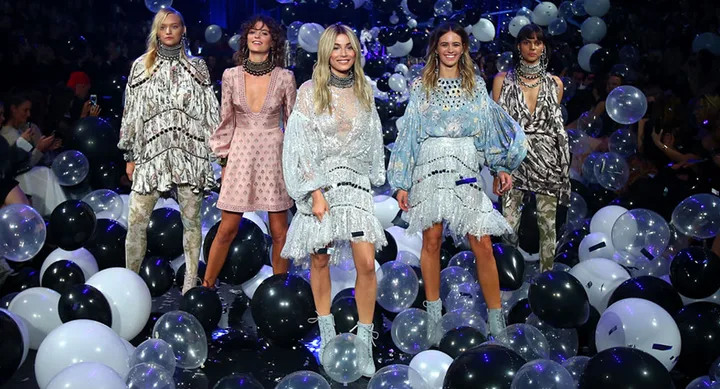 Five models in colorful dresses walk on a runway surrounded by black and white balloons.