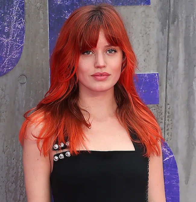 A woman with red hair and bangs, wearing a black outfit with button details, stands against a textured background.