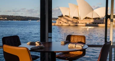 Table set for dining with view of Sydney Opera House over the harbor through large glass windows at sunset.