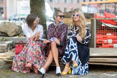 Three women in colorful outfits and sunglasses sit together smiling outdoors on a bench made of pallets.