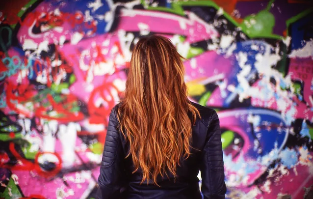 A person with long brown hair wearing a jacket stands in front of a colorful graffiti wall, seen from behind.