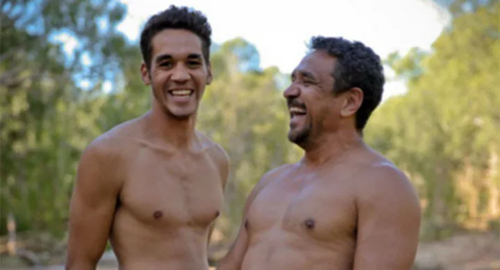 Two shirtless men smiling outdoors, surrounded by trees.