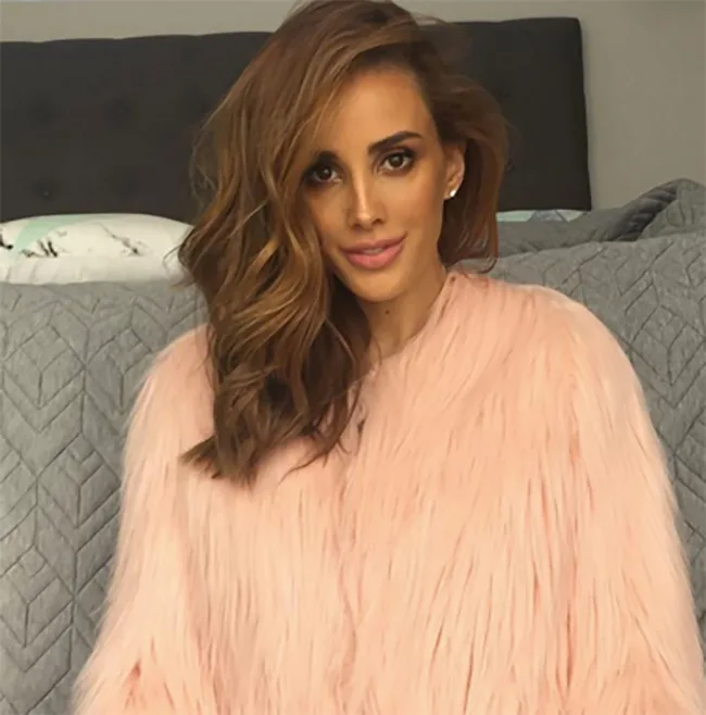 A person with wavy hair wearing a pink faux fur coat sits on a bed, against a patterned gray quilt backdrop.