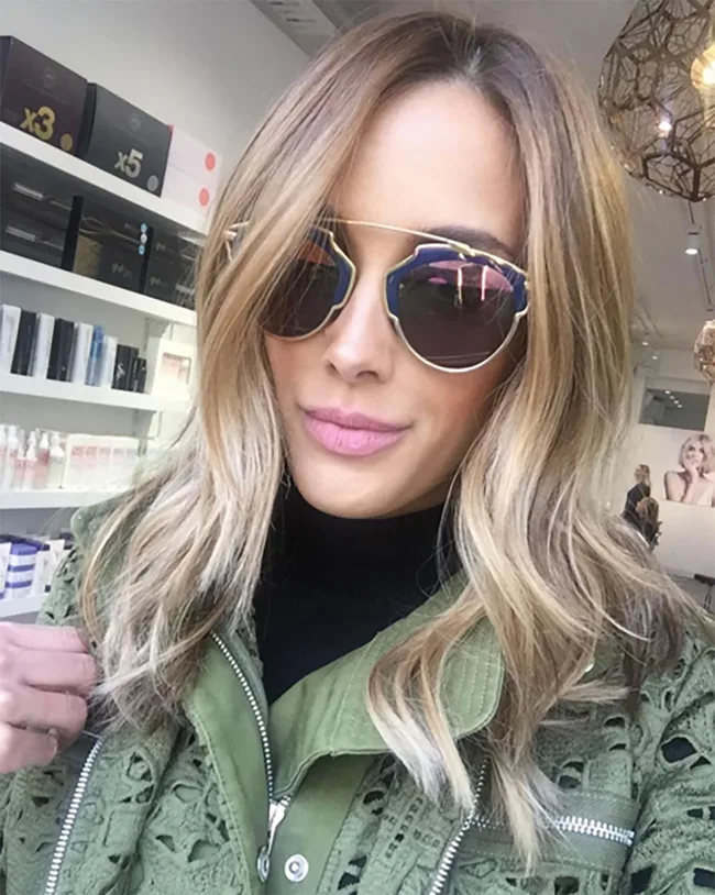 A woman with wavy blonde hair wearing sunglasses and a green jacket inside a store with shelves of products.