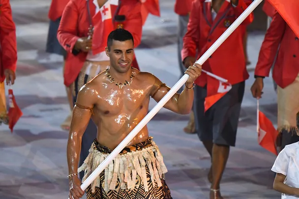 Flag bearer, shirtless in traditional attire, marches with Tongan flag at an event.